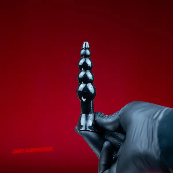 Black latex-gloved hand holding a sleek, graduated pegging plug against a deep red background, representing the psychological and physical aspects of pegging in dominance and submission