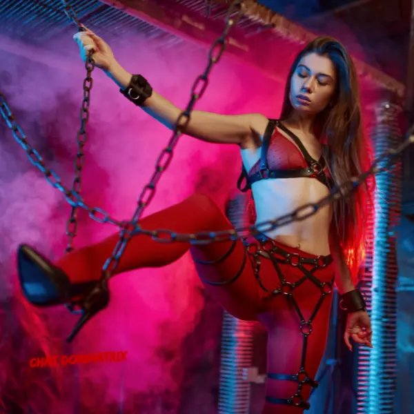 Sensual woman in red lingerie, harnesses, and cuffs posing with chains in a neon-lit industrial setting, symbolizing fetish fashion, bondage, and alternative kink exploration. Featured in 'Kink Exploration' by Aita Goth on ChatDominatrix.com