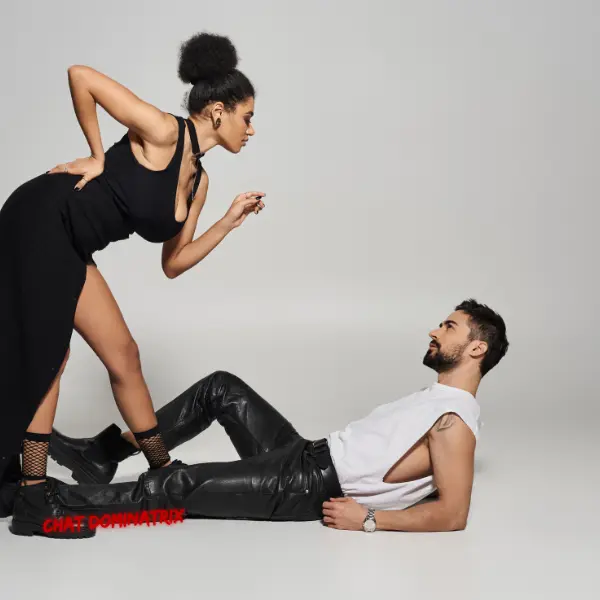 Confident dominant woman in a black dress towering over a submissive man reclining on the floor in leather pants, symbolizing power exchange, obedience, and submissive training in BDSM. Featured in 'Submissive Training: A Guide to Obedience, Devotion & Growth' on ChatDominatrix.com