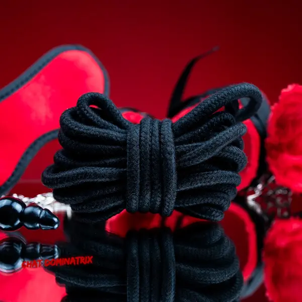 Black BDSM rope neatly coiled with red blindfold, anal beads, and fuzzy handcuffs on a reflective surface, essential beginner bondage toys for BDSM play. Best BDSM toys for beginners guide by Aita Goth on Chat Dominatrix