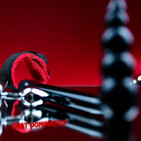 A set of BDSM toys on a glossy black surface with a deep red background, including a red-lined wrist restraint, a metal dildo, and a black anal plug. A moody, sensual setup for Chat Dominatrix’s strap-on and pegging guide in Femdom play